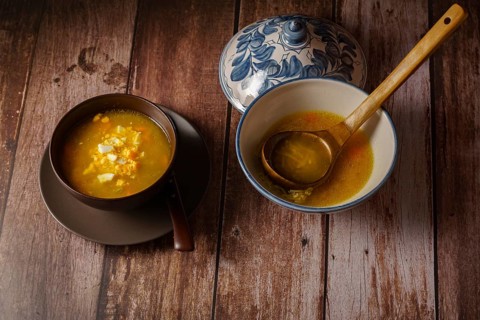Chicken noodle soup in a white soup tureen and a brown bowl with a plate and spoon on a wooden