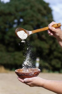 Hands sifting white flour from a wooden spoon into a rustic ceramic bowl, preparing ingredients for