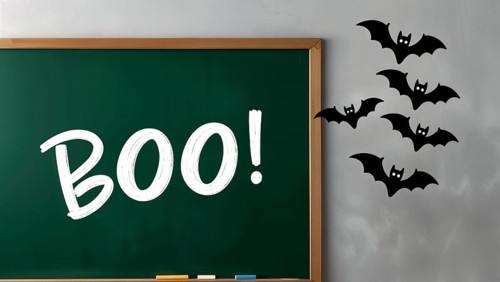 A chalkboard in a classroom shows the word Boo! written in white. Black bats flying against the