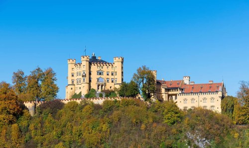 The majestic Hohenschwangau Castle towers over a colorful autumn landscape under clear blue sky,
