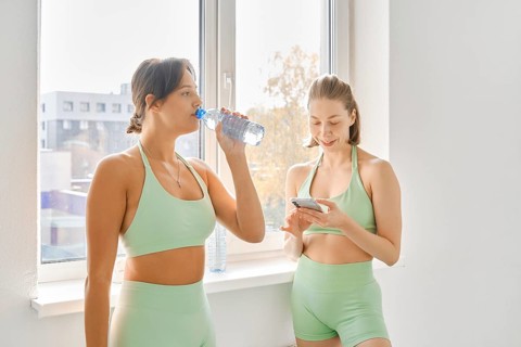 Two women take a break during a fitness stretching class in a bright indoor studio. One drinks