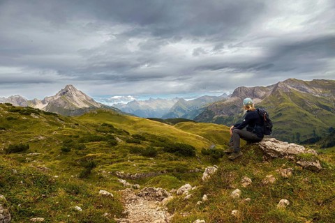 A hiker sits on a hill with a view of a mountainous landscape under a grey cloudy sky, Schröcken,