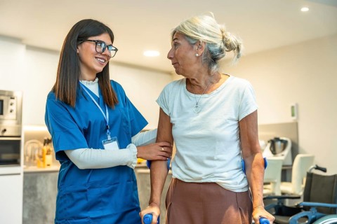 Professional caregiver in uniform supporting an elderly woman using crutches, providing assistance