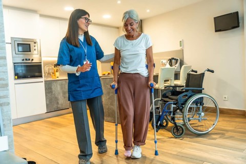 Young female nurse helping a happy elderly woman recovering from injury, walking with crutches