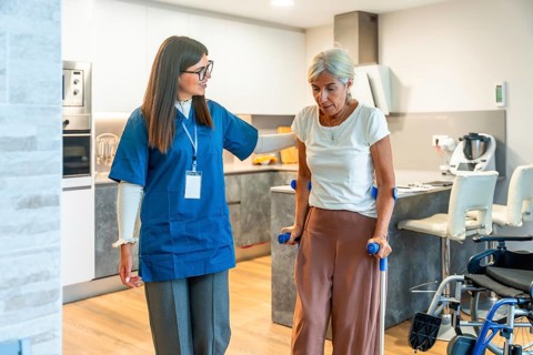 Home care nurse supports elderly woman with crutches during rehabilitation exercises at home,