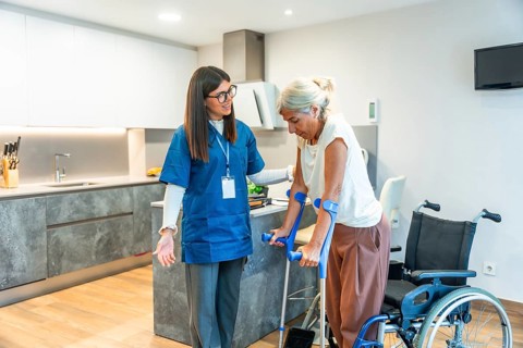 Home nurse assisting a senior woman using crutches to walk, providing support and physical therapy