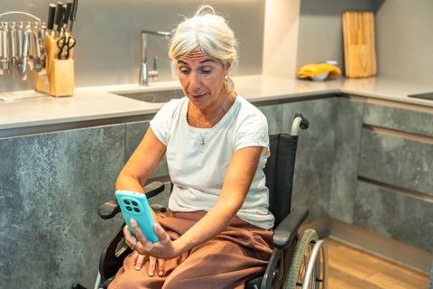 Senior woman in a wheelchair video calling on a smartphone from a modern kitchen, engaging in
