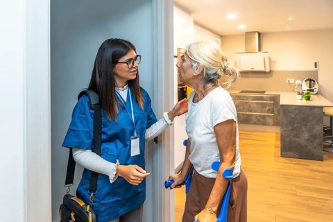 Caregiver wearing scrubs and carrying a medical bag assisting an elderly woman using crutches at