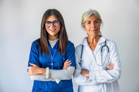 Two women healthcare professionals, a younger nurse in blue scrubs and an older doctor in a white