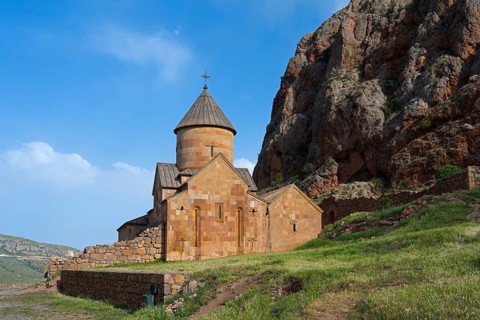A stone monastery building rising on rough rock walls, surrounded by blue skies and green meadows,