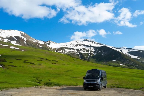 A motorhome stands in front of a green, snow-covered mountain landscape under a blue sky, Camper,