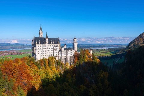Fairytale castle Neuschwanstein on a wooded hill surrounded by autumnal splendor under wide skies,