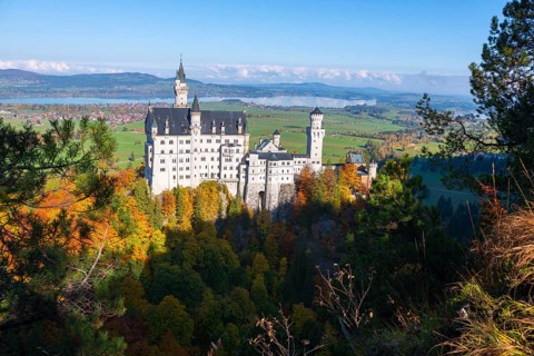 Neuschwanstein Castle on a wooded hill overlooking a green landscape with a wide panorama and blue
