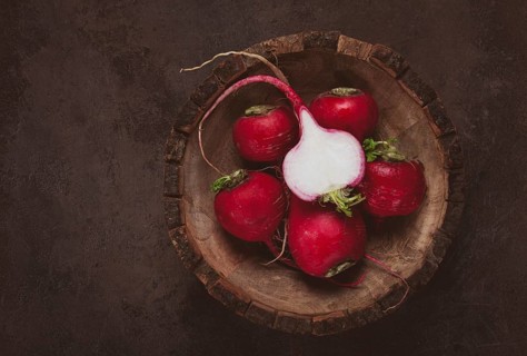 A collection of vibrant red radishes sits in a carved wooden bowl on a dark, textured surface. The