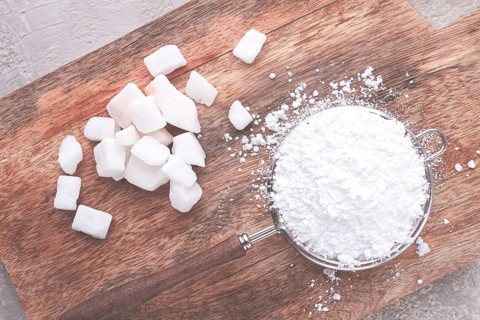 A strainer with powdered sugar, on a wooden cutting board, with pieces of white sugar
