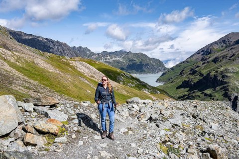 Person posing on rocky path with background of mountains and a lake under blue sky, Lac des Dix