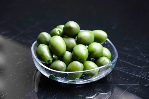 A clear bowl holds a generous pile of green olives. The olives are glossy and vibrant, resting on a