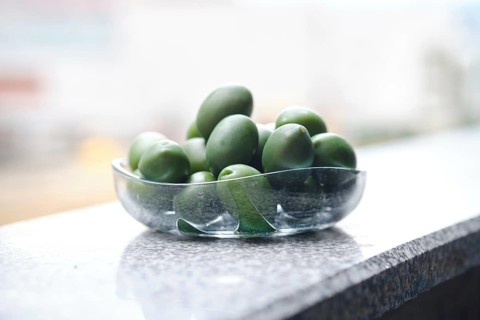 A bowl filled with green olives on a smooth granite surface. The sunlight reflects off the glass