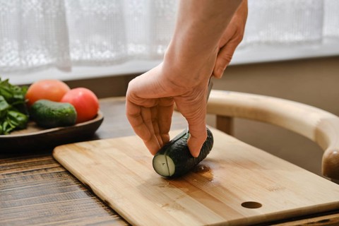 A hand holds a cucumber while cutting it on a wooden board. Fresh tomatoes and greens are arranged