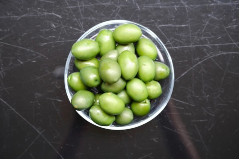 Overhead view of a bowl filled with green olives on a black surface. The olives are vibrant and