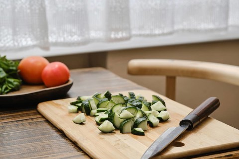 Freshly chopped cucumbers on a wooden cutting board beside a knife. A bowl of tomatoes is visible