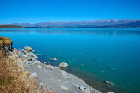 Rocky shore and clear lake with mountain views under clear skies, Lake Pukaki Viewpoint, Pukaki,