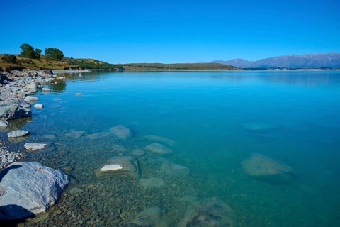 Shore with stones in clear water surrounded by vast countryside and mountains, Lake Pukaki