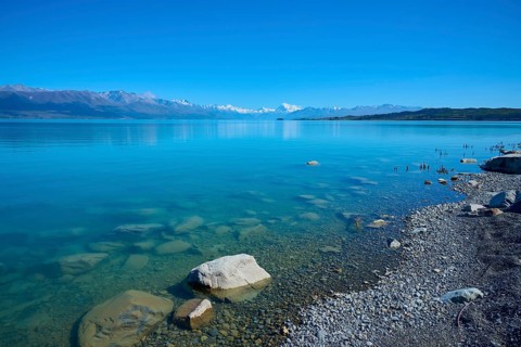 Clear lake with pebble beach and mountain backdrop under blue sky, Lake Pukaki Viewpoint, Pukaki,