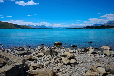 Clear water gently washes against rocky shoreline surrounded by high mountains under bright blue