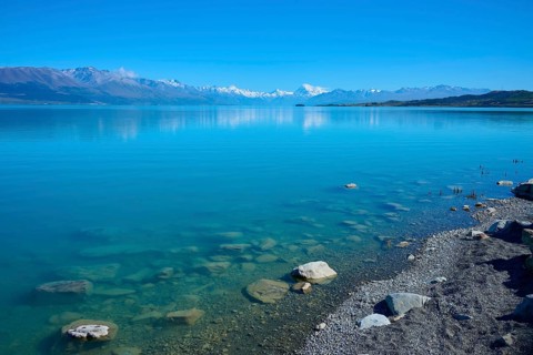 Clear blue water with rocks on the shore and snowy mountains in the background, Lake Pukaki