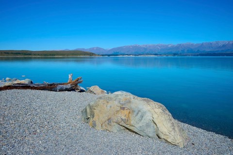 Rocky shore on a calm blue lake with views of distant mountains, Lake Pukaki Viewpoint, Pukaki,