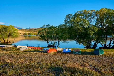 Boats are tipped over on the shores of a calm lake in the evening with wooded hills, Lake McGregor,