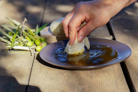 Hand dipping a piece of fresh bread into a shallow plate of extra virgin olive oil, with olives and