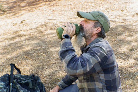 Mature bearded hiker drinking from a canteen during a trekking break in sunlit forest, resting and