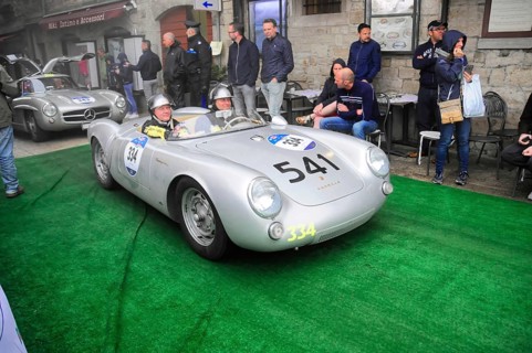 Silver Porsche 550 Spyder with two helmeted drivers drives across the green carpet at the rally,