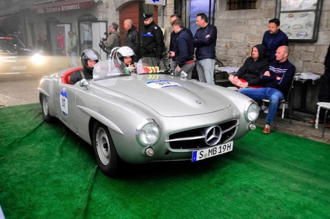 Silver 190 SL Roadster with helmeted occupants slowly across the green carpet, watched by