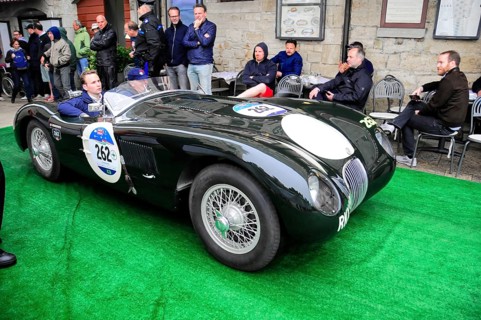 Black jaguar roadster rolls across green carpet during a historic rally, spectators sitting close