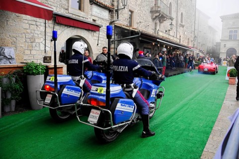 Police motorcycles escort a classic car rally through the stone old town, audience lines the green
