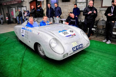 Aluminum-colored racing roadster with blue drivers at a vintage car rally on green carpet, purist