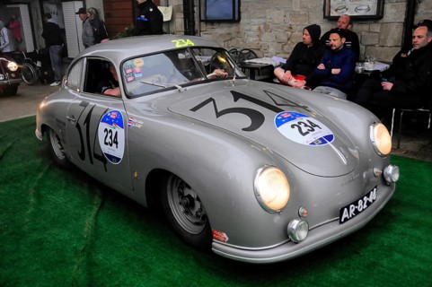 Silver Porsche Coupé rolls across green carpet in front of spectators on a stone wall at a historic