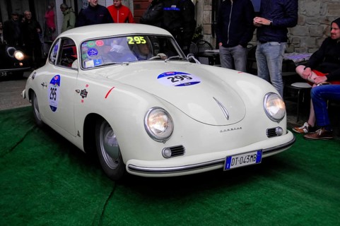 White Porsche 356 with glowing headlights drives across green carpet during a vintage car rally,