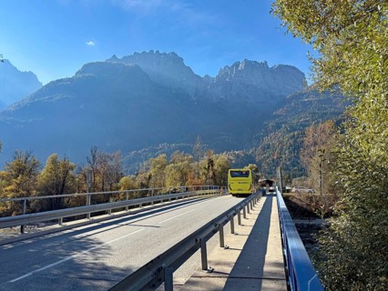 A yellow bus crosses a bridge under a blue sky, surrounded by autumnal mountains, Lienz Dolomitem,