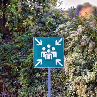 Green outdoor sign, pictogram showing group of people surrounded by four arrows, escape sign for