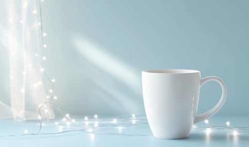 A white coffee cup sits on a table next to a string of lights. The lights are twinkling and