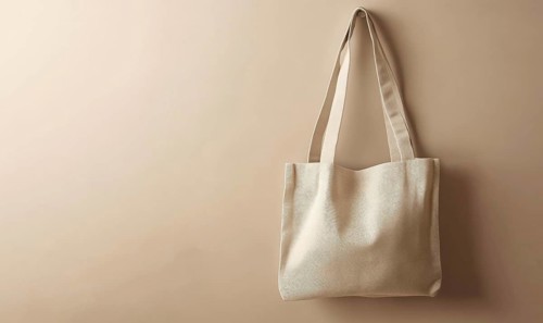 A tan tote bag is hanging on a wall. The bag is made of a light material and has a simple design.