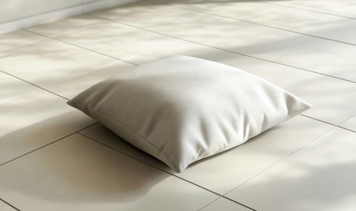A white pillow sits on a tile floor. The pillow is small and square in shape. The floor is white