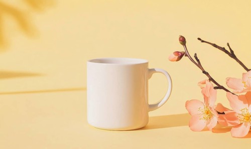 A white coffee cup sits on a yellow background with pink flowers in the foreground. The cup is