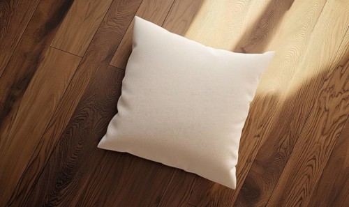 A white pillow sits on a wooden floor. The pillow is placed in the center of the image, and the