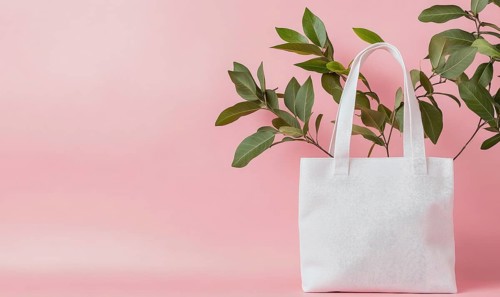 A white bag with a leaf on it is on a pink background. The bag is placed in front of a plant, which