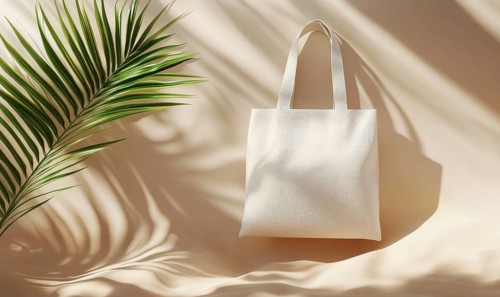 A white tote bag is sitting on a tan surface with a leafy green palm tree in the background.